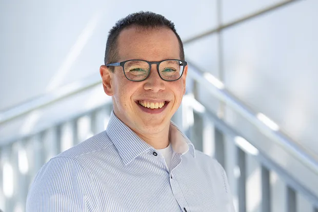 Smiling man with glasses and striped shirt, standing in front of modern architecture.