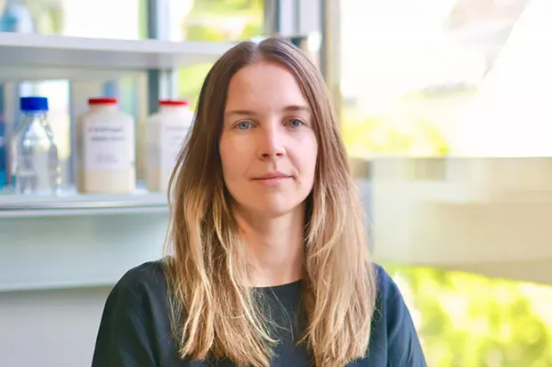 Woman with long, light hair stands in front of a window, with bottles and containers visible in the background.
