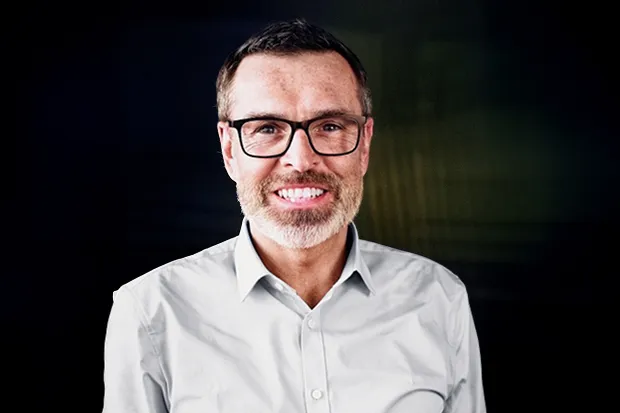 Man with glasses and beard, smiling against a dark background, wearing a light-colored shirt.