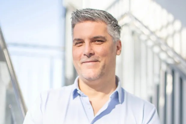 Man with gray hair, smiling, wearing a light-colored shirt, standing in a modern, light-filled room.