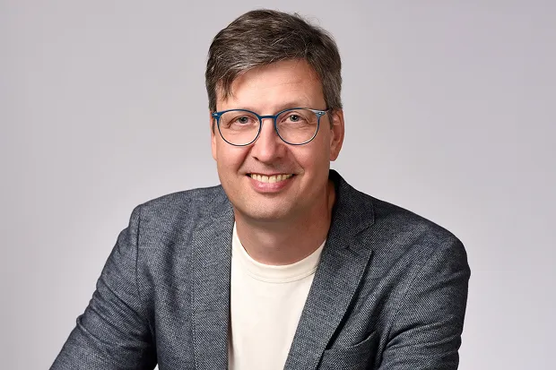 Smiling man wearing glasses, wearing a gray blazer and light-colored T-shirt, standing in front of a neutral background.
