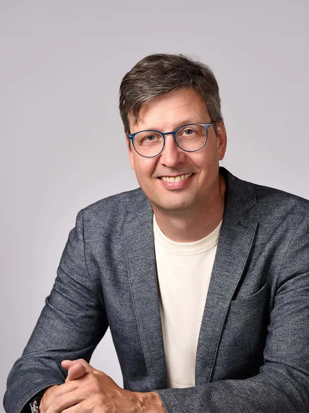 Man wearing glasses and a gray blazer, smiling and sitting with his hands folded in front of a neutral background.