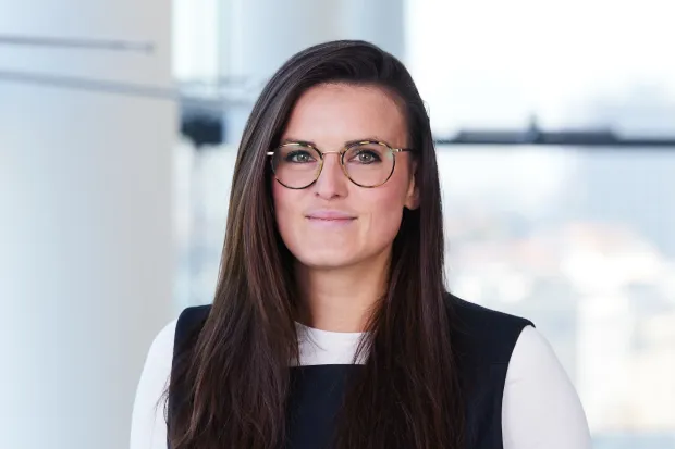 Woman with long, dark hair and glasses smiles in front of a bright background with windows.
