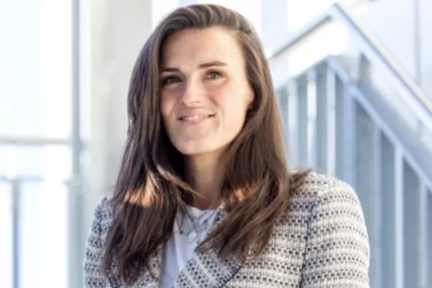 Woman with long, dark hair and glasses smiles in front of a bright background with windows.