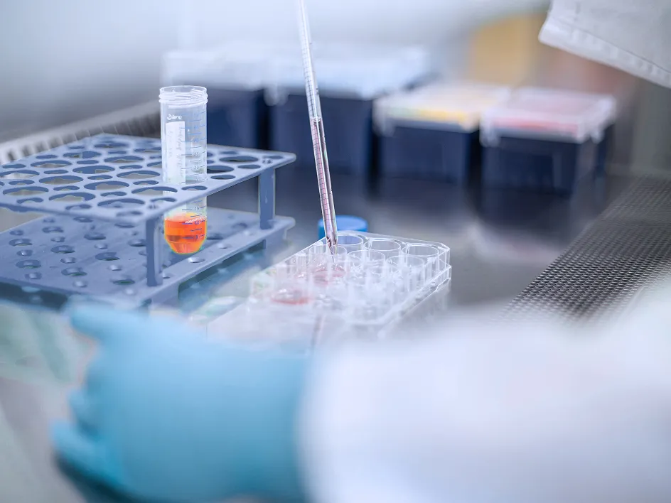 Laboratory scene with a gloved hand handling test tubes and a pipette, surrounded by storage containers.