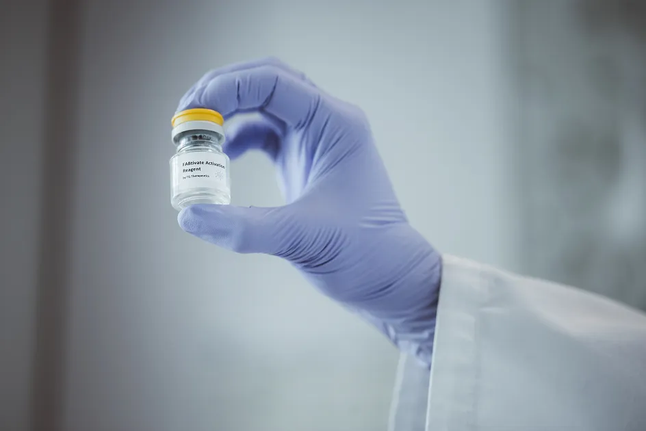 A hand wearing a purple glove is holding a small bottle with a yellow cap.