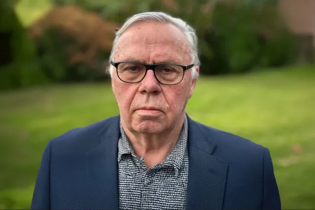 An older man with glasses and a serious expression is wearing a blue blazer and standing in front of a green meadow.