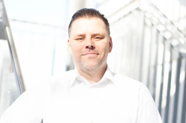 Man in a white shirt smiles in front of a bright, blurred background scene.