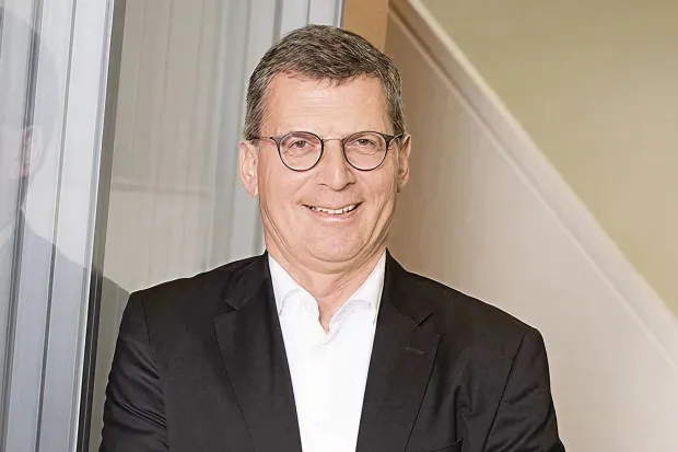 Man in a suit with glasses smiles in front of a staircase and a glass wall.