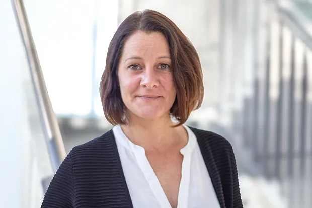Woman with shoulder-length brown hair standing in front of a staircase in a bright room. She is wearing a white top and a black jacket.