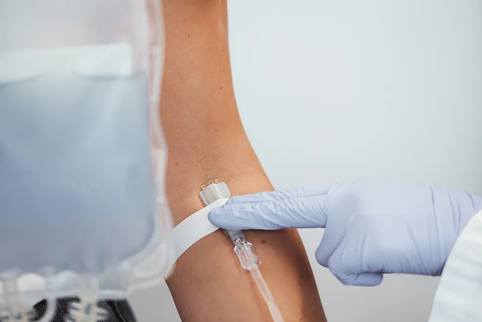A hand wearing a blue glove points to an intravenous line in an arm, while an IV bag is visible in the foreground.