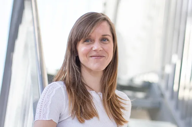 Woman with long brown hair and a smiling face, standing in a bright, modern room.