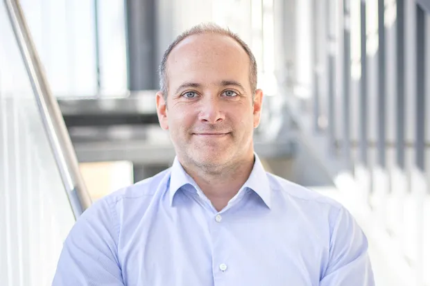 Man with short hair, smiling, wearing a light blue shirt, sitting in front of a modern staircase.