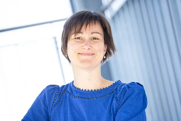 Woman with short hair wearing a blue blouse smiles in front of a bright, modern background.