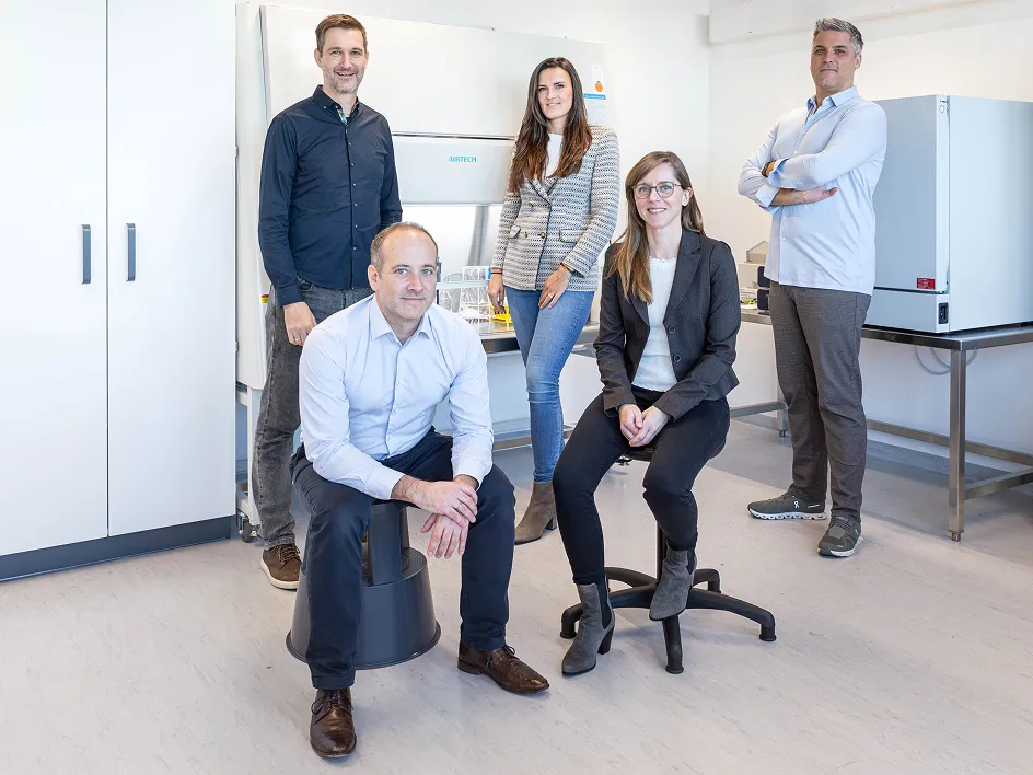 Five people are standing and sitting in a modern laboratory, surrounded by equipment and white walls.