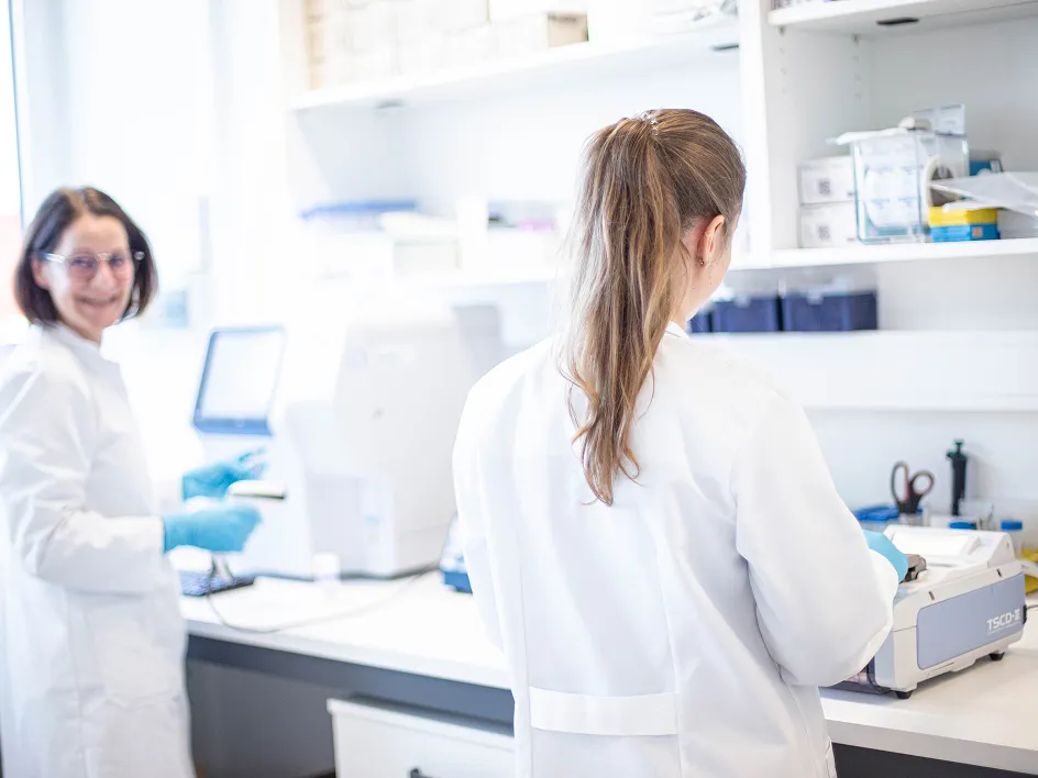 Two women in lab coats are working in a bright laboratory; one is smiling while the other is standing next to a piece of equipment.