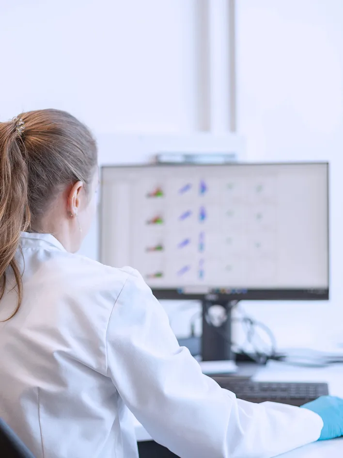 Woman in lab clothing sitting in front of a computer screen displaying scientific graphics.