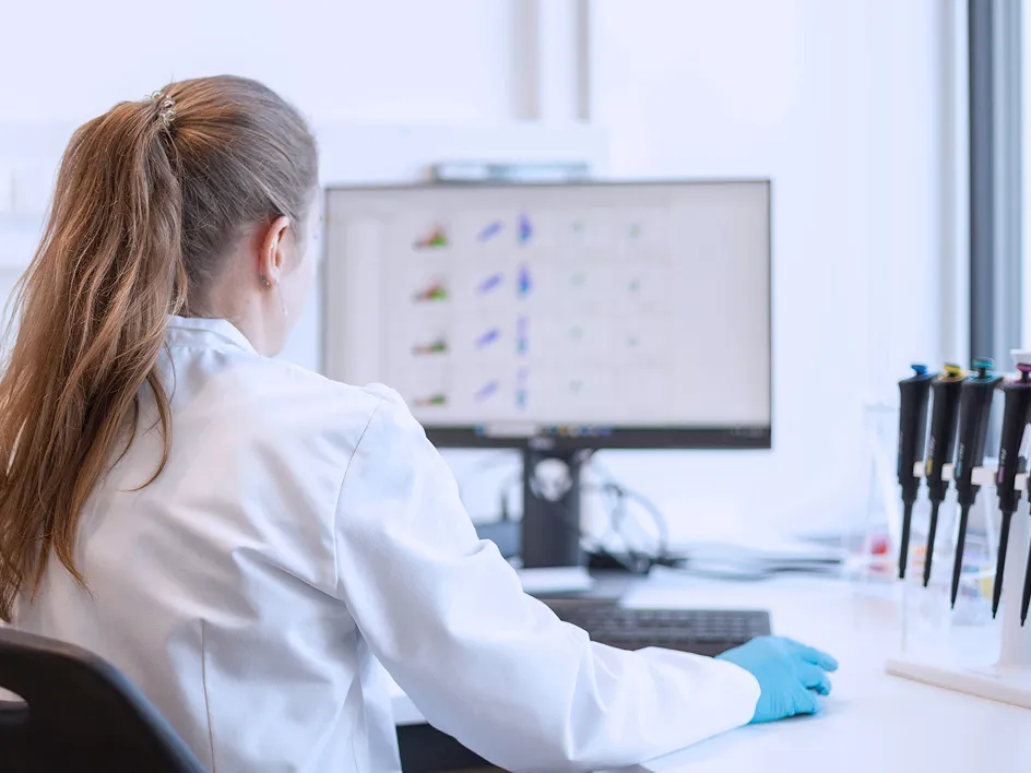 A person wearing a lab coat sits at a desk and looks at a computer screen displaying scientific data.